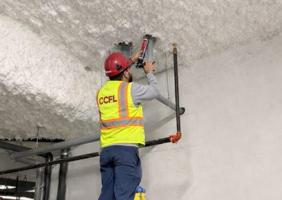 An installer in a high-visibility vest applying firestop sealant with a caulking gun around a pipe through a rated wall.