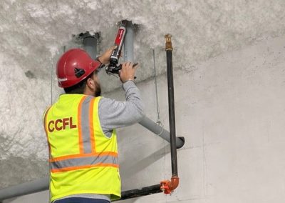 An installer in a high-visibility vest sealing a pipe penetration through a concrete wall with firestop sealant.